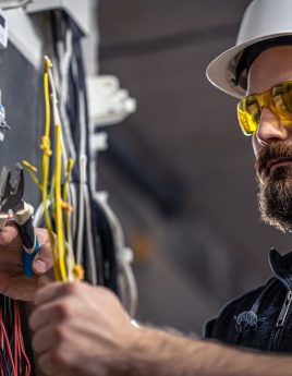 A male electrician works in a switchboard with an electrical connecting cable. Male electrician at the checkout counter on a blurred background of a switchboard.